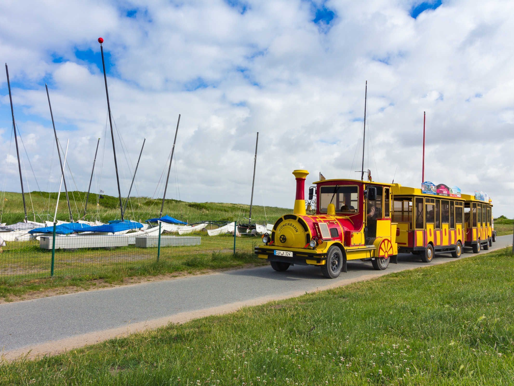 A yellow and red train with "DUNENBAHN" written on it, moving along a road with a grassy area and boats in the background.