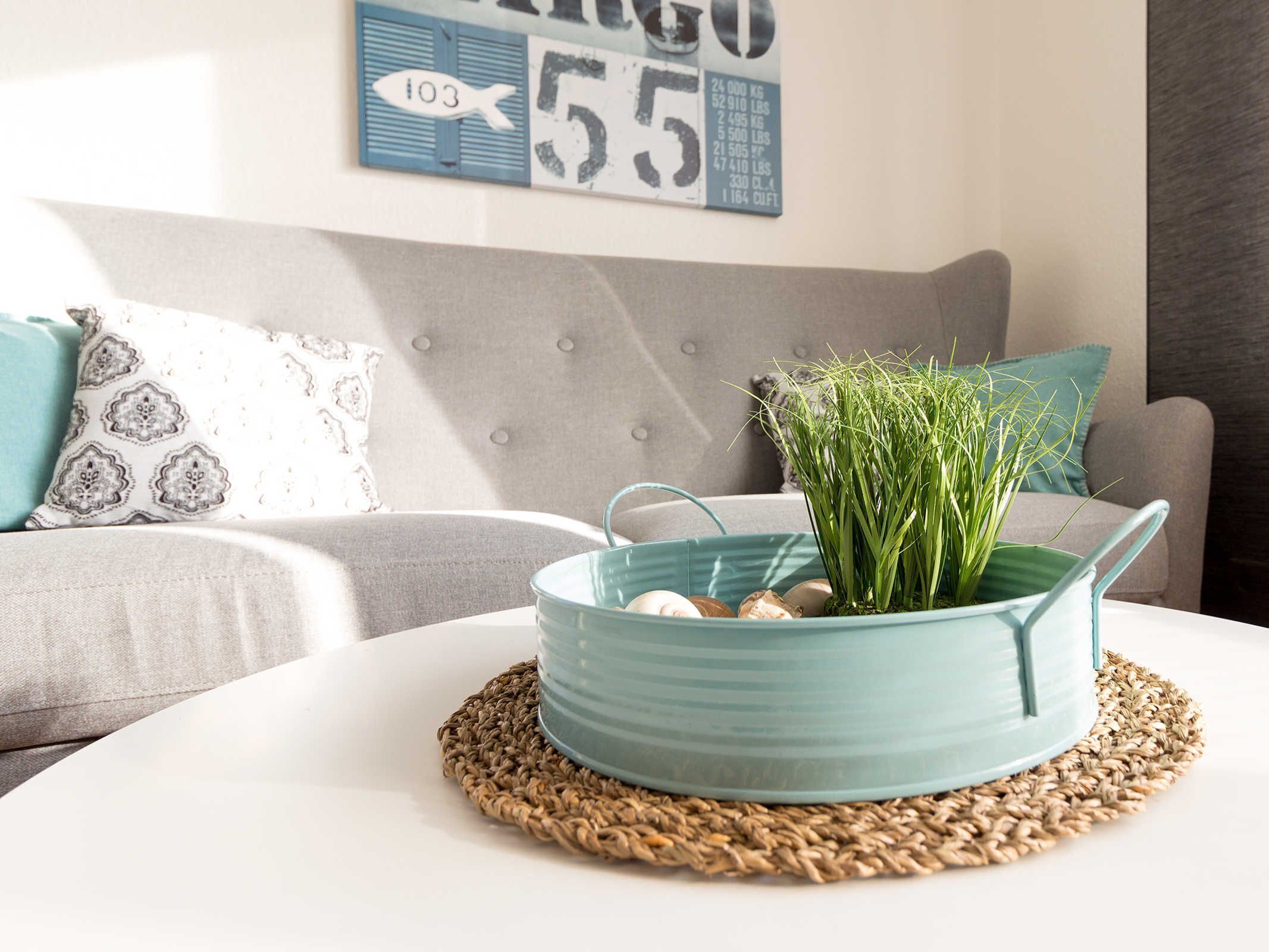Ein Wohnzimmer mit einem türkisfarbenen Topf auf einem weißen Tisch, einem grauen Sofa mit gemusterten Kissen und einer Wand mit einem Hinweisschild.