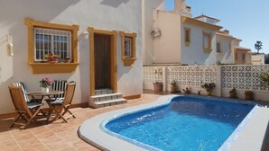 Une piscine avec une terrasse carrelée, une table entourée de chaises et des plantes en pot.