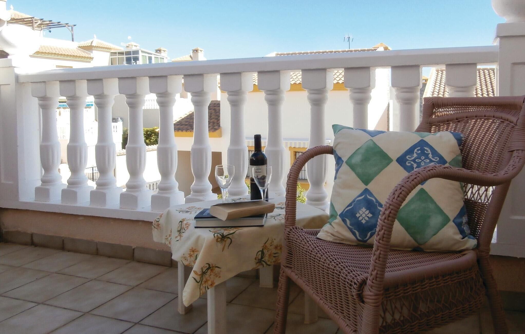 Una silla de mimbre con un cojín estampado en un balcón con baranda blanca, una mesa con mantel floral y una botella de vino.