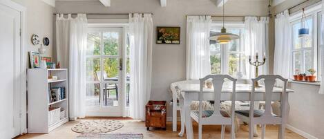 A bright, airy dining room with white furniture, a large window with curtains, and a view of greenery outside.