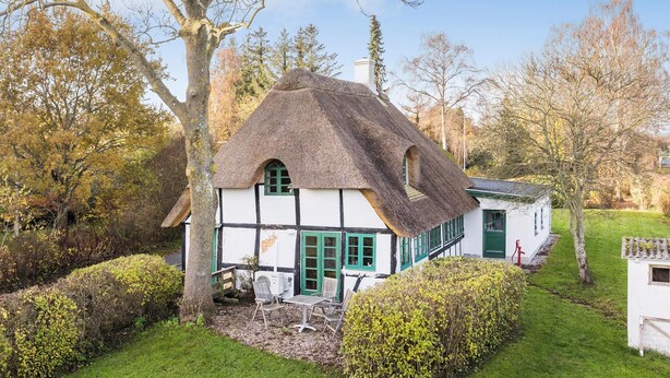 Une maison traditionnelle à toit de chaume, aux murs blancs et aux volets verts, entourée d’un jardin bien entretenu et d’arbres.