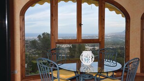 Ein Essbereich mit Tisch und Stühlen, einer dekorativen Vase und Blick auf eine Küstenlandschaft durch ein großes Rundbogenfenster.