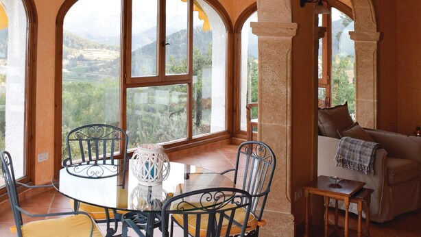 Ein Zimmer mit einem großen Bogenfenster, das einen Blick auf eine gebirgige Landschaft bietet. Es gibt einen runden Glastisch mit Stühlen, einen kleinen Holztisch und eine Couch mit Überwurfdecke.