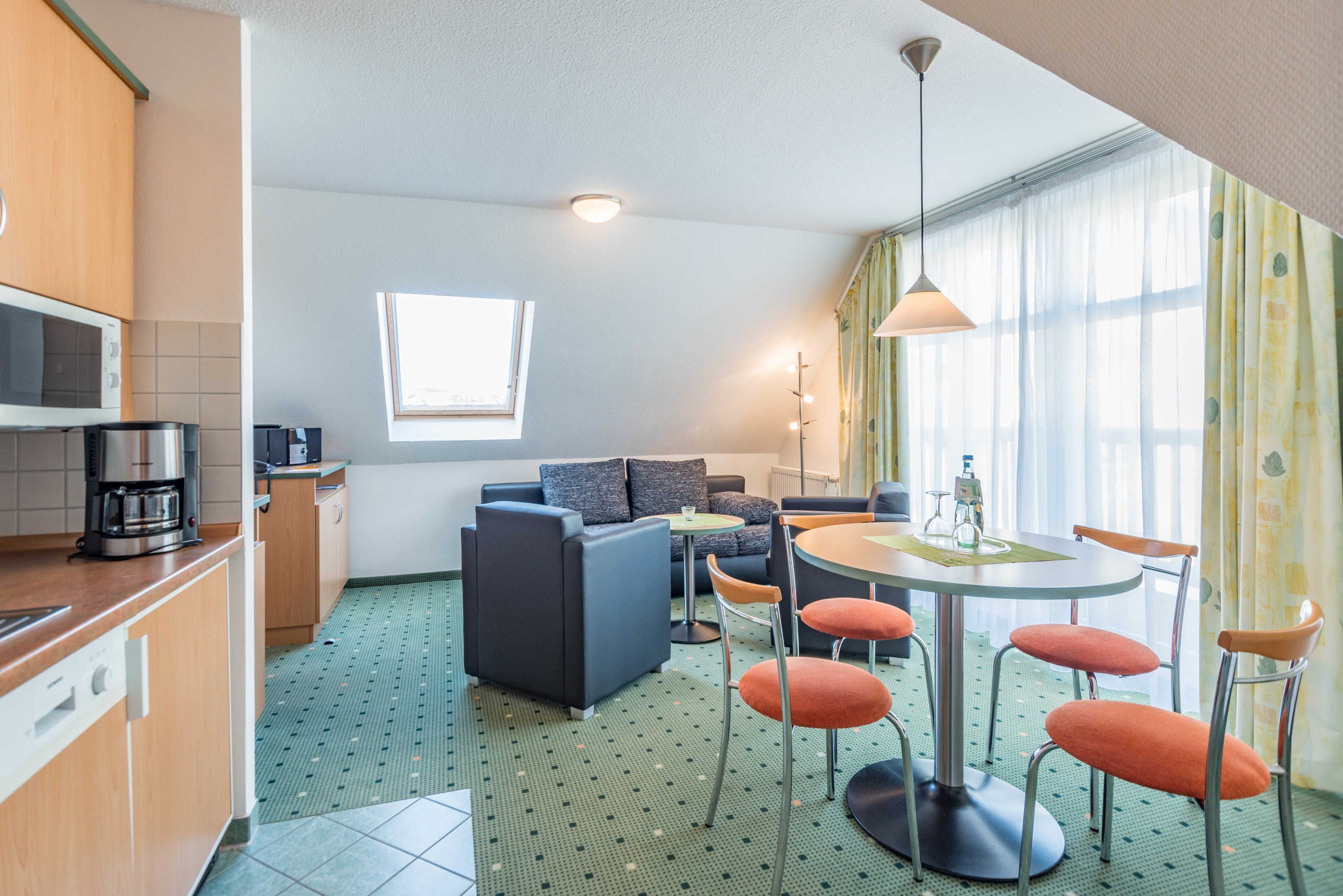Een moderne hotelkamer met een kitchenette, een bank, een eettafel met stoelen en een groot raam met gordijnen.
