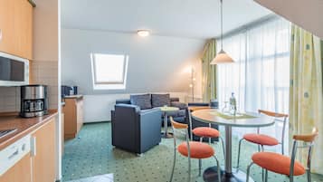 Een moderne hotelkamer met een kitchenette, een bank, een eettafel met stoelen en een groot raam met gordijnen.