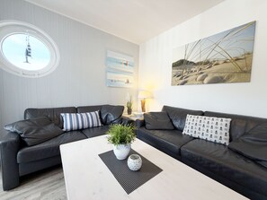 Un salon moderne avec deux canapés en cuir noir, une fenêtre ronde, une table basse blanche et un tableau représentant une scène de plage.