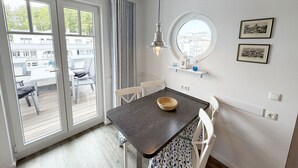 Un coin repas avec une table en bois foncé, des chaises blanches, un luminaire suspendu, une fenêtre ronde et une vue sur les bâtiments à l’extérieur.