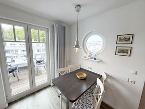 A dining area with a dark wooden table, white chairs, a hanging light fixture, a round window, and a view of buildings outside. - MR: Villa Rosa Whg. 16 Sea magic with 2 roof terraces - Villa Rosa Whg. 16 (Sellin)