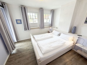 A bedroom with a double bed, a bedside table with a lamp, a nightstand, and a window with curtains. - MR: Villa Rosa Whg. 16 Sea magic with 2 roof terraces - Villa Rosa Whg. 16 (Sellin)