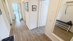 Un couloir doté d’un plancher en bois, de murs blancs et de portes donnant sur d’autres pièces. Des tableaux encadrés ornent les murs, et un banc avec un radiateur se trouve dans un coin.