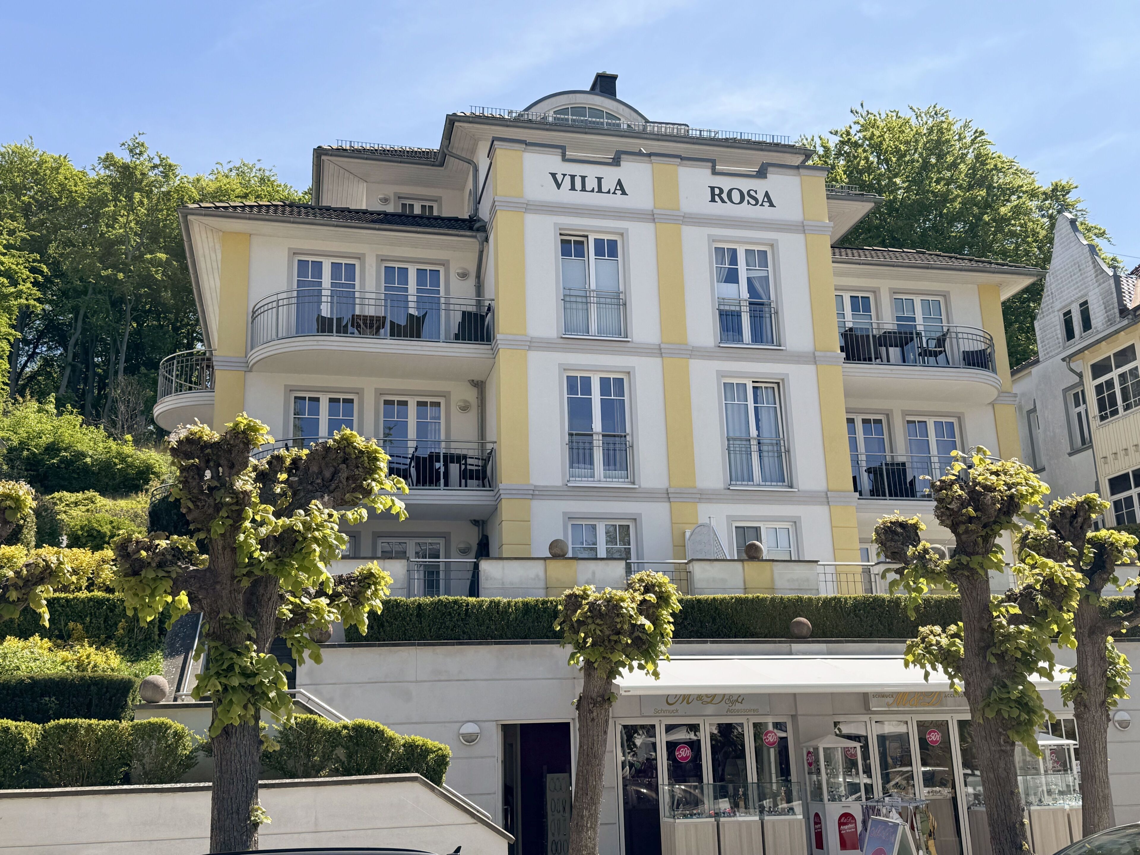 A multi-story building named "Villa Rosa" with balconies, a storefront, and well-maintained greenery.