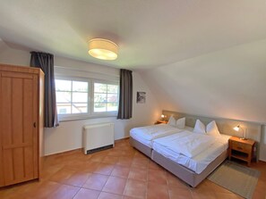 Quarto com uma cama, mesa de cabeceira, radiador, janela com cortinas e uma porta.