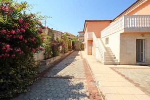 Stroll through a charming Mediterranean street lined with flowering bushes and trees, surrounded by residential buildings with light-colored walls and balconies.