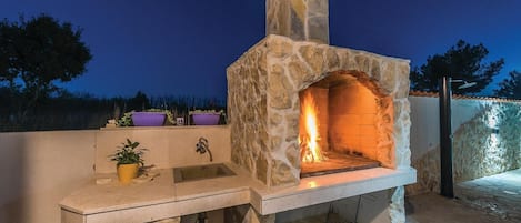 Horno de piedra para pizzas al aire libre, con fuego encendido en su interior, rodeado de plantas en macetas y un lavamanos.