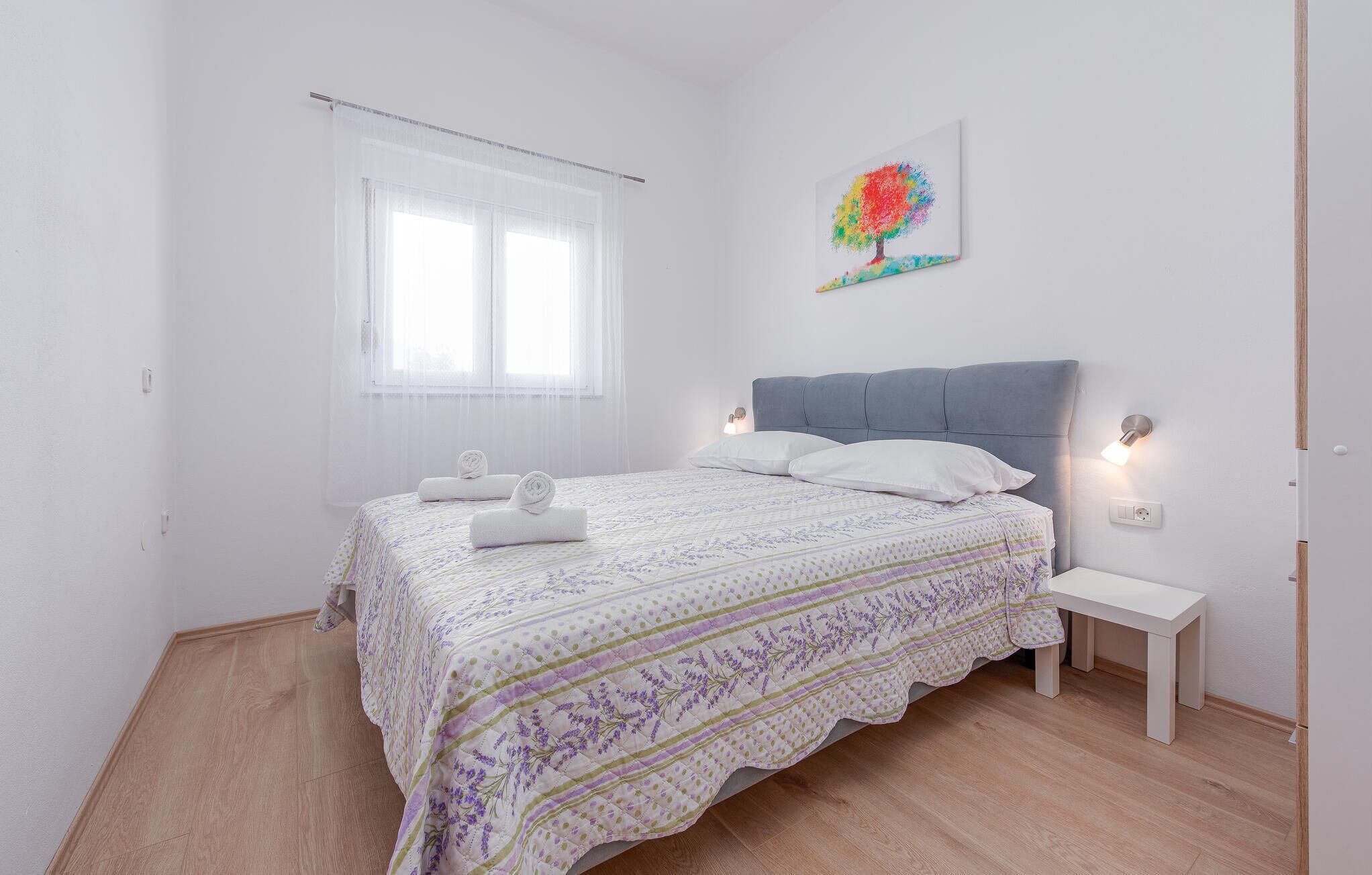 Une chambre à coucher comprenant un lit, une table de chevet, une lampe fixée au mur et une fenêtre ornée de rideaux légers.
