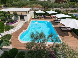 Une piscine rectangulaire dotée d’un jacuzzi intégré, entourée d’une terrasse en bois et de parasols blancs.