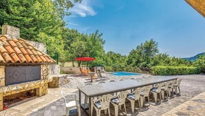 Une terrasse extérieure avec un barbecue en pierre, une piscine et un coin repas avec des chaises et une table.