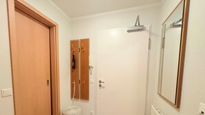 Un corridoio con una porta in legno, una porta bianca, uno sgabello di vimini, un termosifone bianco e uno specchio appeso al muro.
