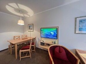 Une pièce comprenant une table à manger en bois, quatre chaises, un téléviseur posé sur un meuble, un ventilateur de plafond et deux tableaux encadrés accrochés au mur.