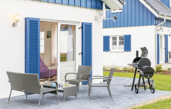 An outdoor patio with wicker chairs, a barbecue grill, and a sliding glass door leading to a room with blue shutters.