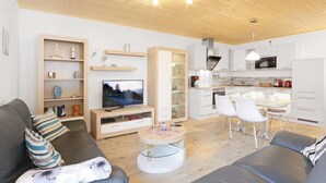 A modern living room with a grey sofa, a coffee table, and a TV. There is a kitchen area with white cabinets and a dining table with chairs.