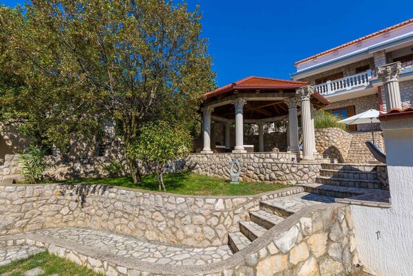 A stone-walled garden with a gazebo, steps leading to a building with columns, and a clear blue sky.