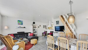 A modern living room with a black sofa, wicker chairs, a dining table with chairs, a TV, and a staircase.