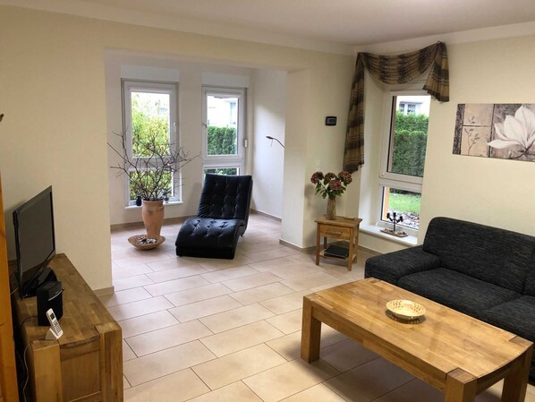 Ein Wohnzimmer mit einer Couch, einem Holztisch, einem schwarzen Ledersessel, einem Fernseher und einem Blumentopf.