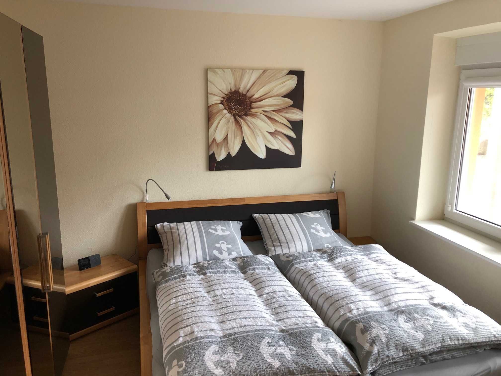 A bedroom with a bed, a nightstand, a lamp, a window, and a wall-mounted flower painting.
