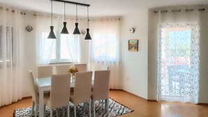 Zona pranzo con tavolo e sedie, lampadari a sospensione, tappeto decorato e finestra con tende leggere.