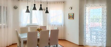 Un comedor con una mesa y sillas, lámparas colgantes, un tapete estampado y un ventanal con cortinas translúcidas.