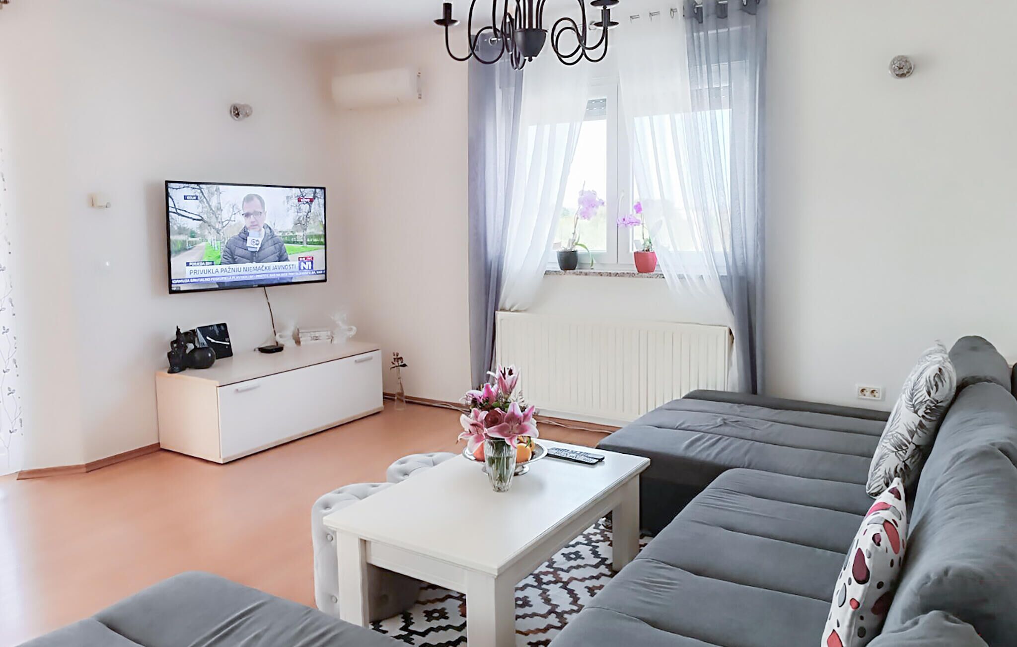 A modern living room with a flat-screen TV, a white sideboard, a grey sofa, and a small white coffee table with a vase.
