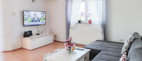 A modern living room with a flat-screen TV, a white sideboard, a grey sofa, and a small white coffee table with a vase.