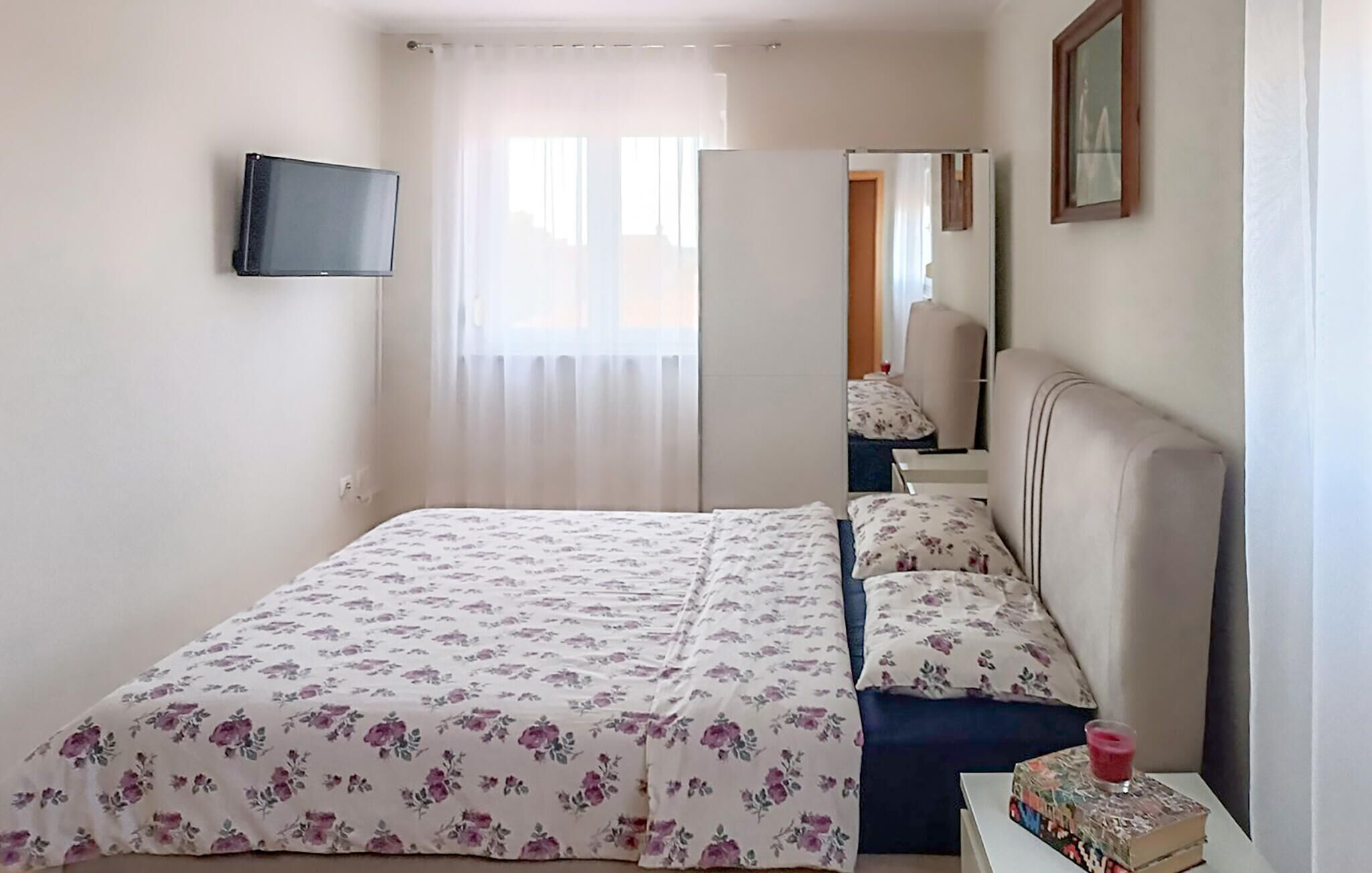 Een slaapkamer met een bed, nachtkastje, spiegel en een aan de muur bevestigde televisie.