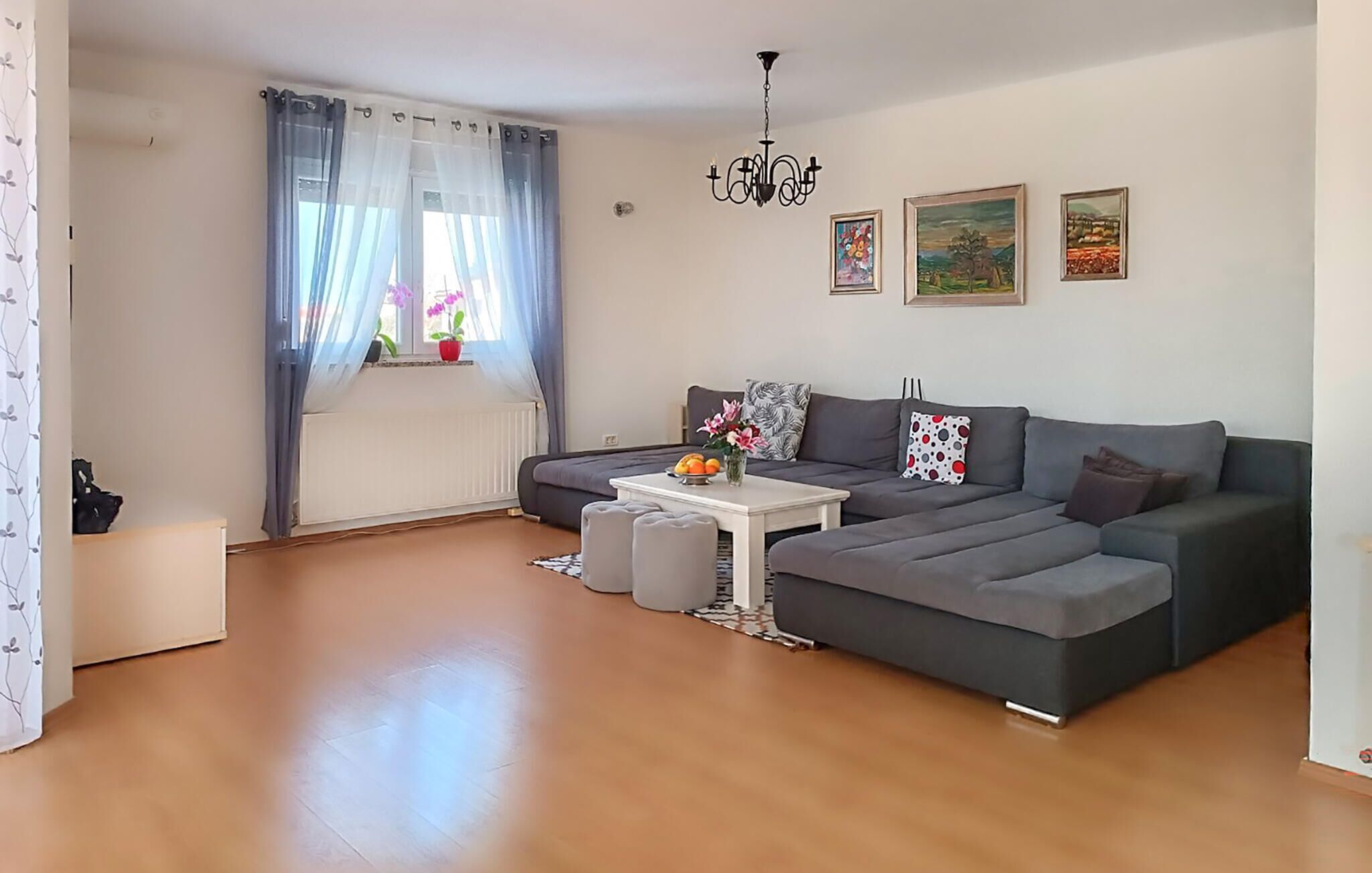Un salon moderne avec un canapé d’angle gris, une table basse blanche et une fenêtre ornée de rideaux légers.