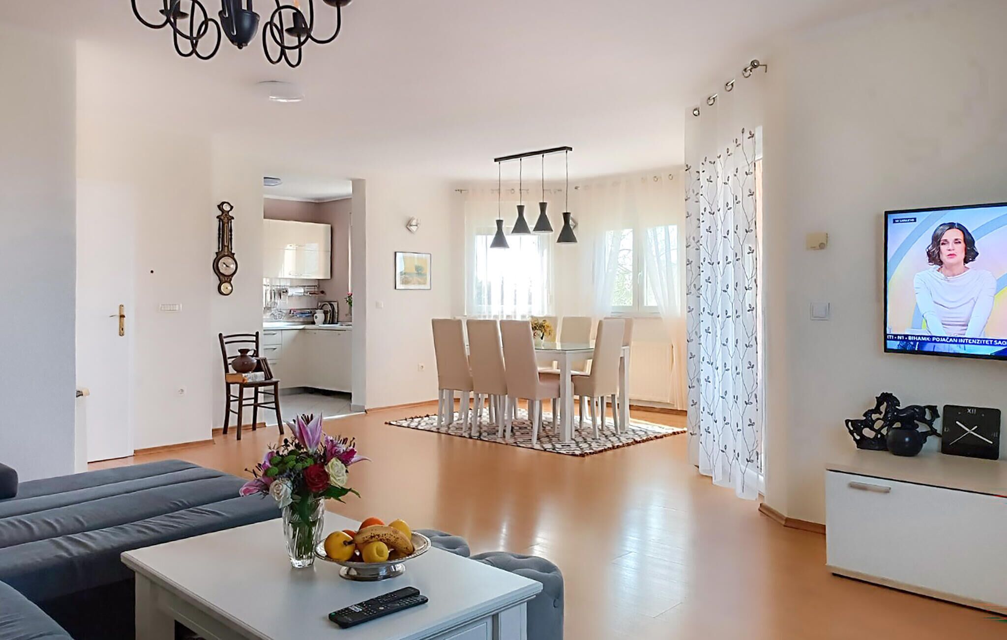 Un salon comprenant un canapé, une table basse avec un vase et un panier de fruits, un téléviseur, ainsi qu’un coin repas avec des chaises et une table.