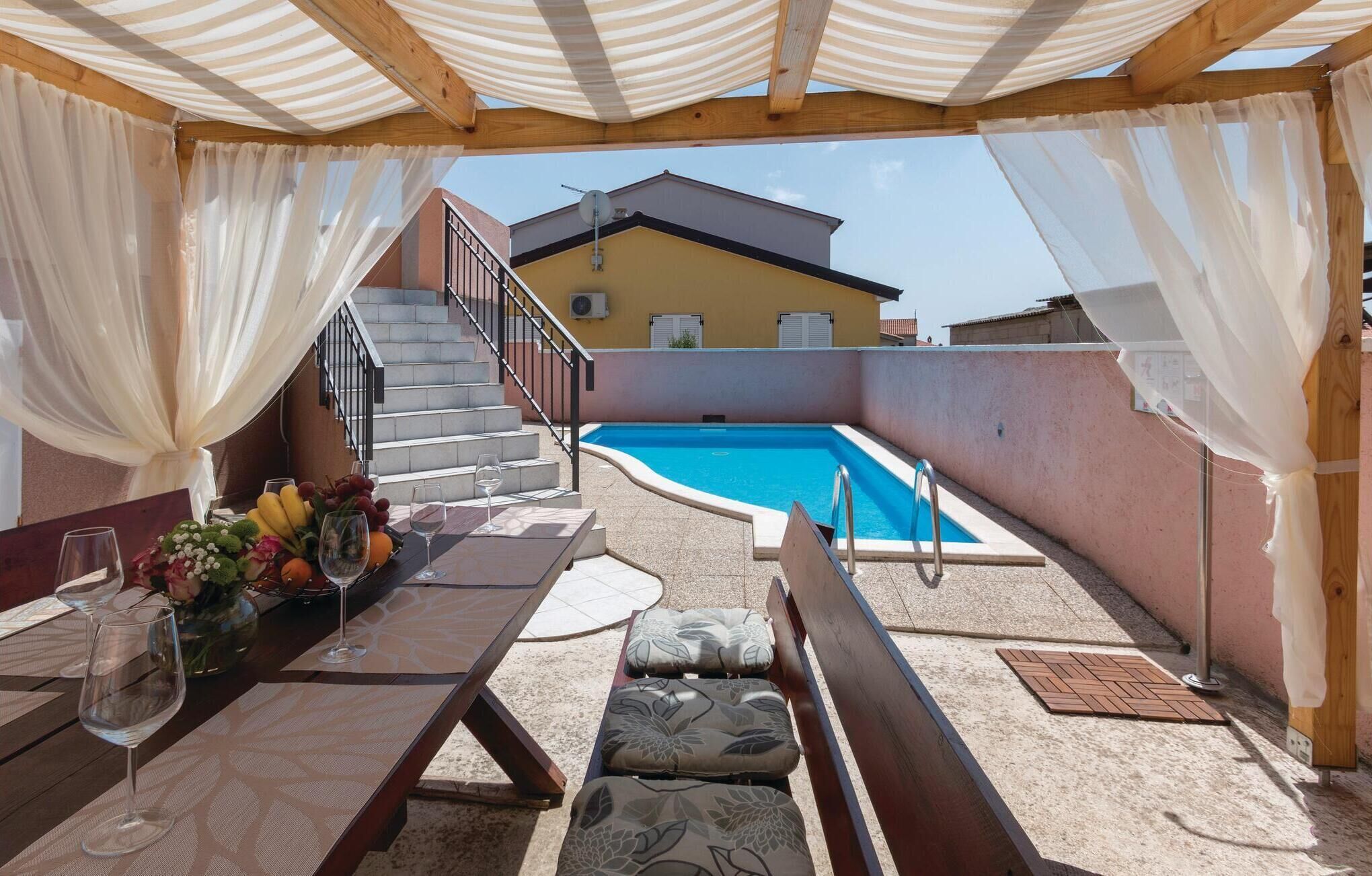 Zona de piscina al aire libre con un porche de madera, una mesa con copas de vino y fruta, y una escalera que conduce a un edificio.