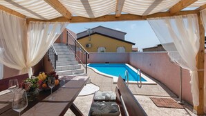 Area piscina esterna con un pergolato in legno, un tavolo con bicchieri da vino e frutta, e una scalinata che conduce a un edificio.