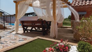 Un'area salotto esterna coperta con una panca e un tavolo, circondata da una piscina e un giardino.