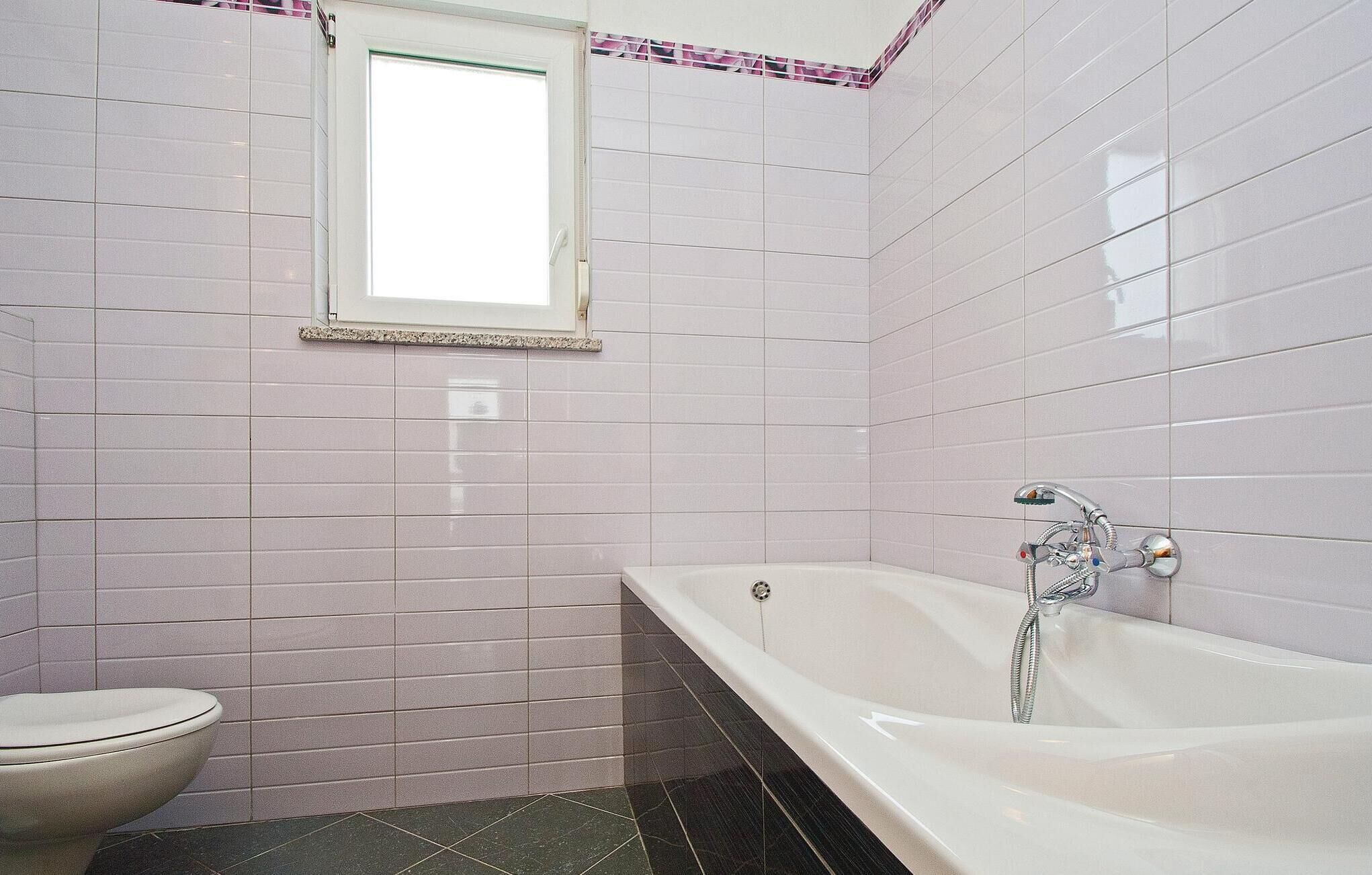 A bathroom with white tiled walls, a window, a toilet, and a bathtub with a showerhead.