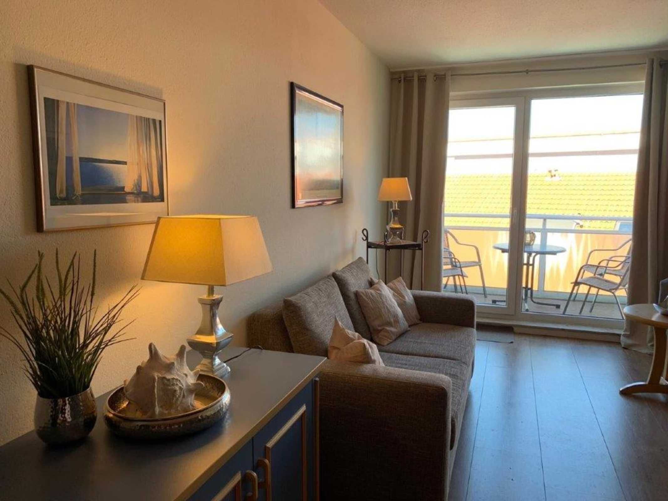 Ett vardagsrum med en soffa, ett soffbord, en lampa och en växt. På väggen hänger två inramade tavlor, och det finns en balkong med stolar och ett bord.