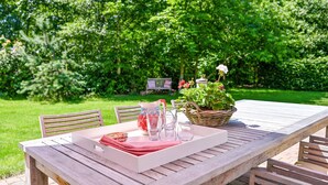 Une table en bois surmontée d’un plateau blanc, un panier en osier rempli de fleurs et un banc dans un jardin verdoyant.