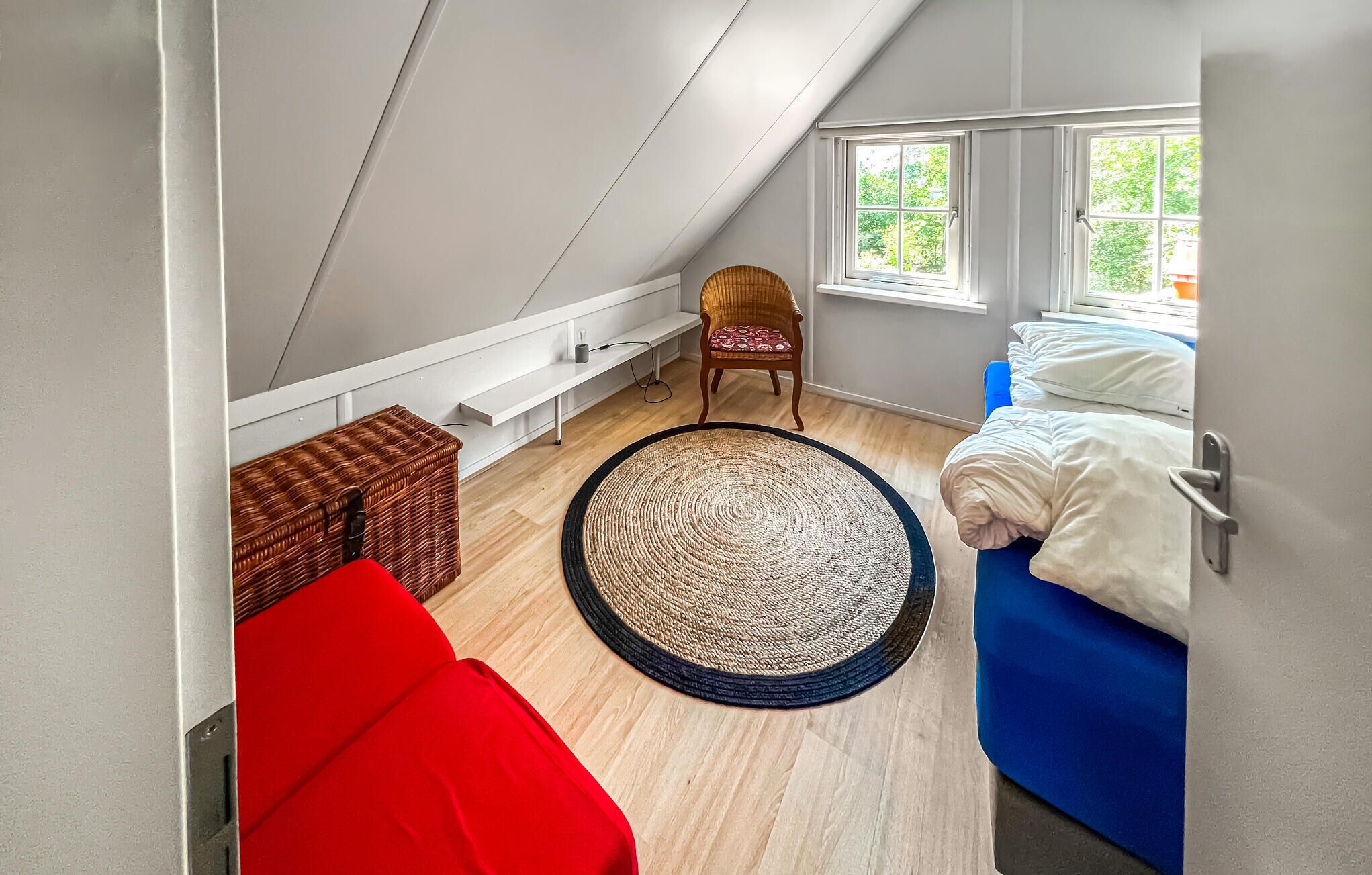 Een kleine, gezellige kamer met schuine plafonds, een houten vloer, een rode bank, een blauw bed, een rieten stoel en een groot geweven vloerkleed.