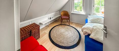 Een kleine, gezellige kamer met schuine plafonds, een houten vloer, een rode bank, een blauw bed, een rieten stoel en een groot geweven vloerkleed.