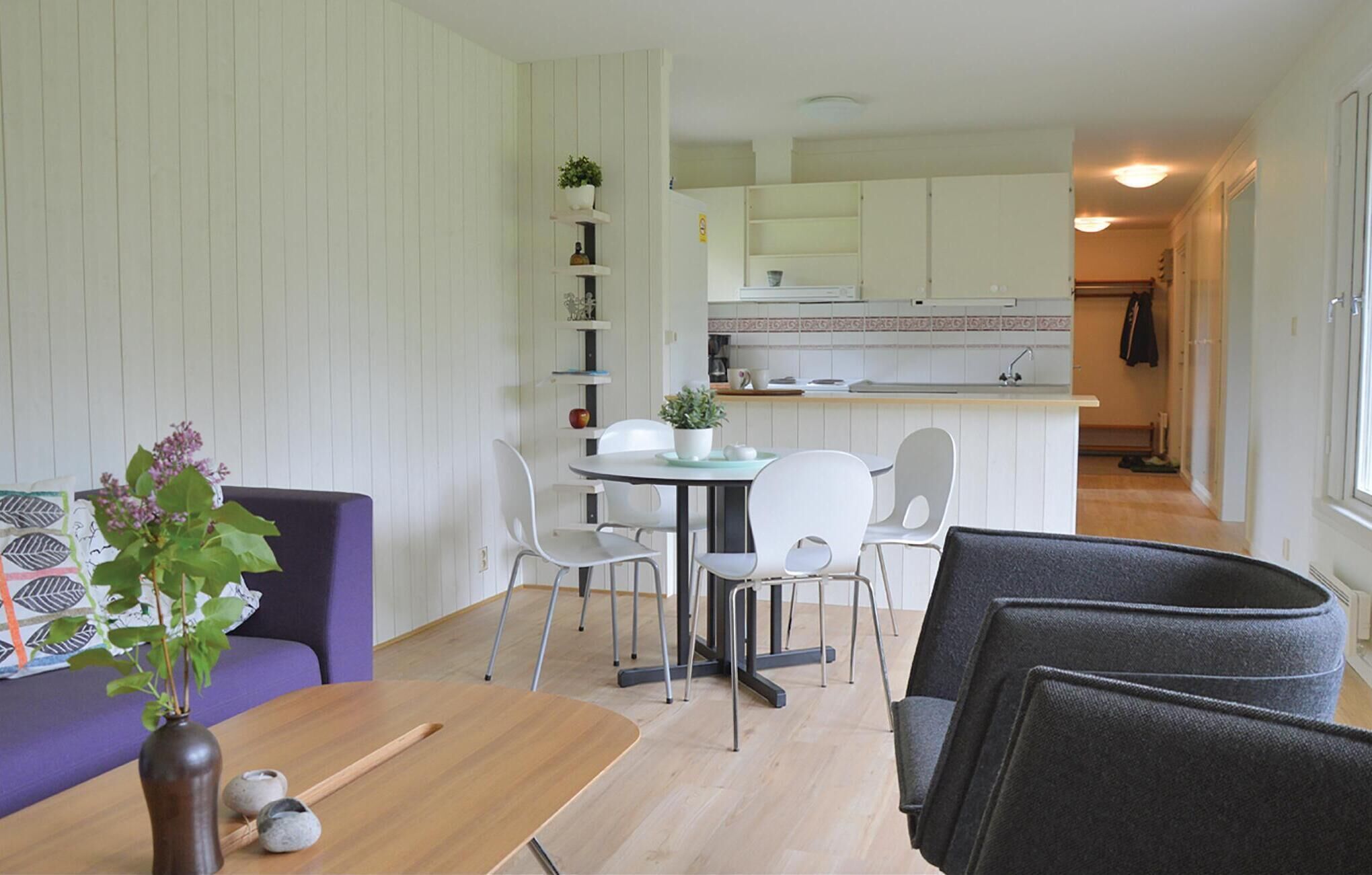 Un salon moderne avec une table en bois, un canapé violet et des chaises blanches. On aperçoit, au fond, une cuisine avec des meubles blancs et un évier.