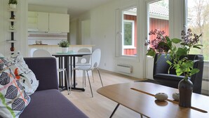 A modern living room with a sofa, a dining table with chairs, a kitchen area, and a large window with a view of trees.