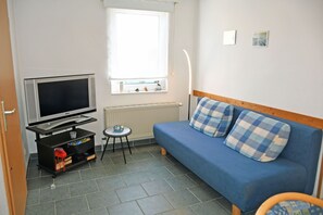 A small room with a blue sofa, a television on a stand, a small table, and a window with blinds.