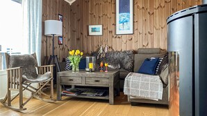Ein gemütliches Wohnzimmer mit Holzboden, einer Couch, einem Schaukelstuhl, einem Couchtisch mit Büchern und einer Blumenvase, einer Lampe und einem Kamin.