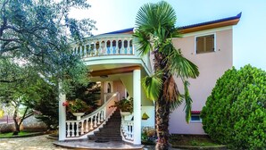 Una casa de dos pisos con un balcón curvo, una escalera que lleva al piso superior y un jardín bien cuidado con diversas plantas y una palmera.